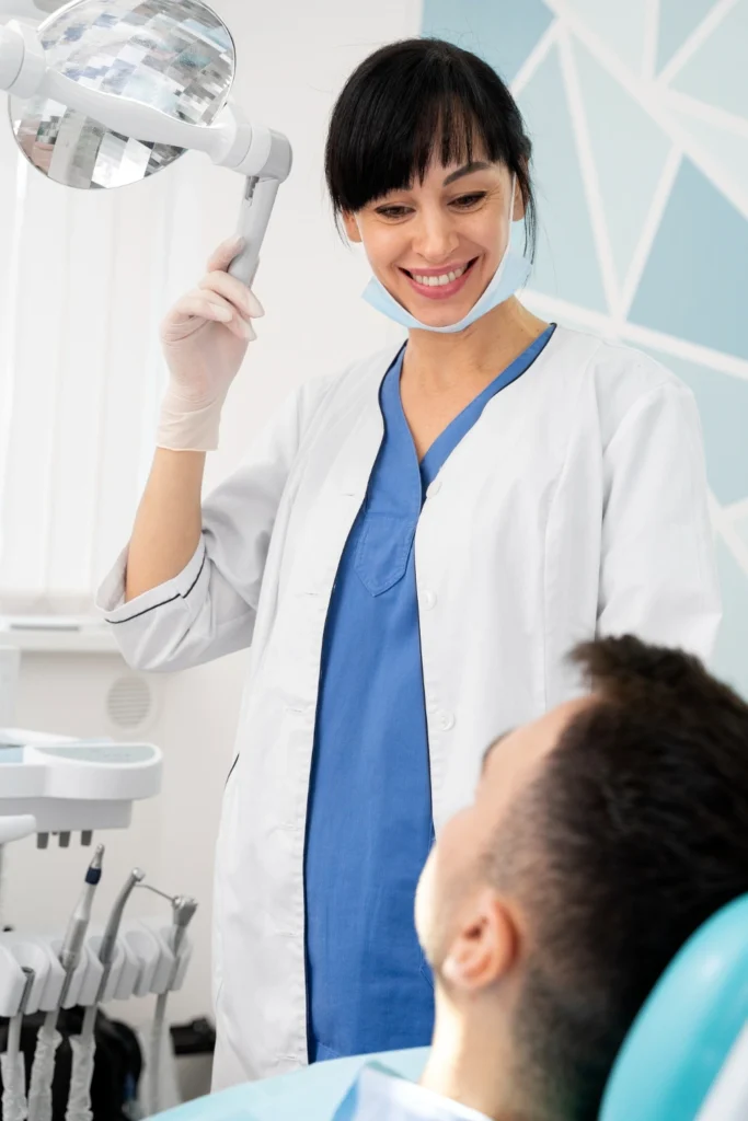 A professional clinical photograph showing advanced patient care. A smiling female clinician in blue scrubs and a white lab coat adjusts a modern overhead surgical lamp while looking down at a seated male patient. This image represents the advanced clinical technology, meticulous diagnostic illumination, and personalized surgical care delivered at icónica dental group.