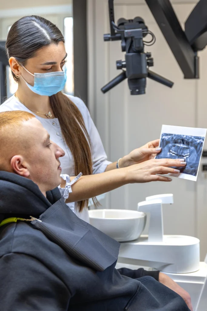 A professional clinical photograph showing an advanced diagnostic consultation. A female clinician wearing a blue face mask holds a panoramic dental x-ray print for a seated male patient, with a precision surgical dental microscope visible in the background. This image represents the digital radiography diagnostics, advanced microscope-enhanced dentistry, and transparent treatment consultations delivered at icónica dental group.
