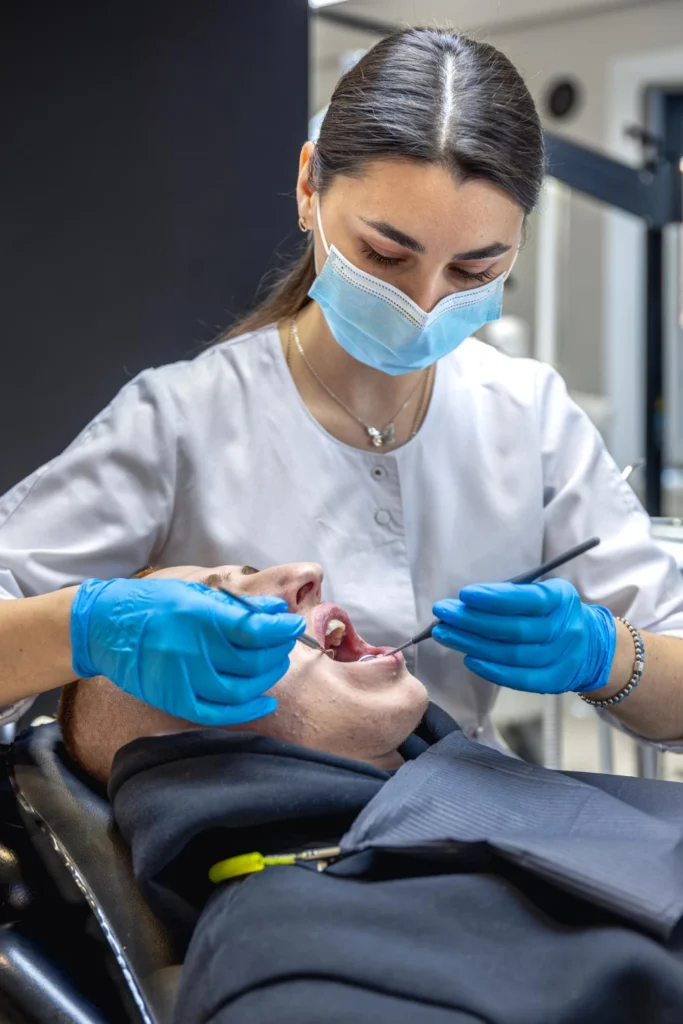 A professional photograph showing a female clinician wearing a white medical coat, surgical face mask, and blue protective gloves to perform an intraoral surgical procedure. She uses precise diagnostic tools to evaluate the sublingual structural tissues of a male patient. This image represents the advanced laser frenectomies, specialized mandibular tori removals, and precise soft tissue oral surgeries performed at icónica dental group.