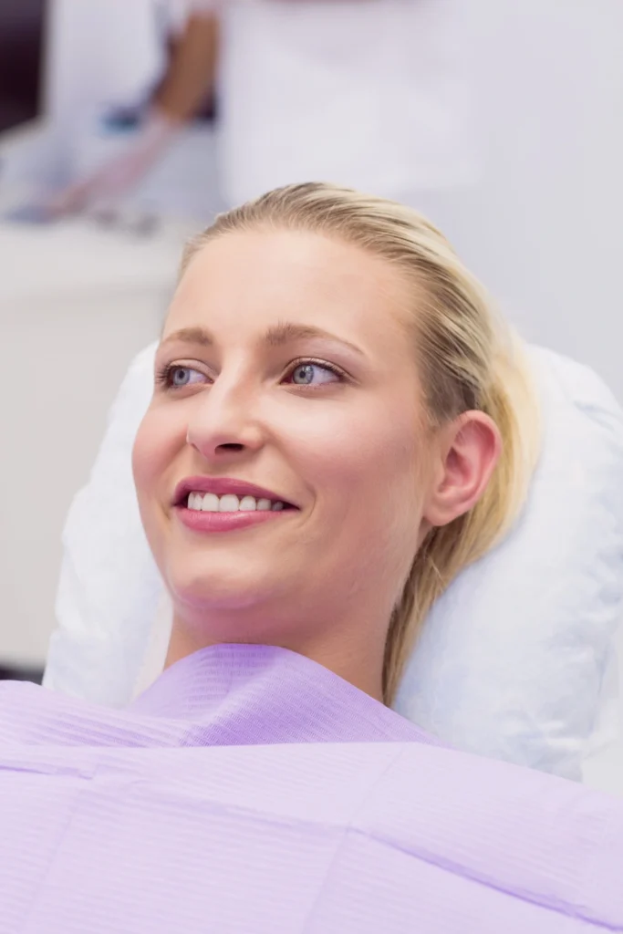 A professional photograph showing a relaxed, smiling blonde female patient resting comfortably in a dental chair under a lavender protective bib. This image represents the high-end patient experiences, customized comfort menu transformations, and complete peace of mind delivered during All-on-X full-arch dental implant procedures at icónica dental group.