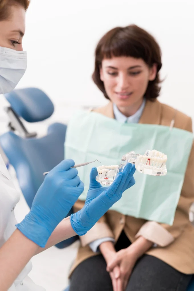 A professional photograph showing a female clinician wearing a white coat, protective face mask, and blue gloves holding a clear anatomical model of a jaw. She utilizes a dental explorer tool to explain structural bone architecture during a consultation with a female patient. This image represents the autogenous bone grafting, advanced pre-prosthetic surgical planning, and hard tissue ridge reconstruction practiced at icónica dental group.