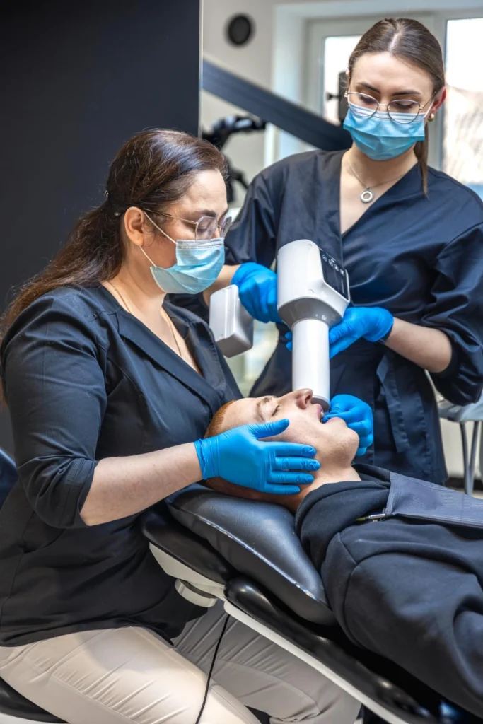 A professional photograph showing two female clinicians wearing dark scrubs, protective face masks, and blue gloves preparing a male patient in a treatment chair. One clinician stabilizes the patient while the other operates a handheld portable intraoral dental x-ray machine. This image represents the precise bone grafting diagnostics, comprehensive sinus lift mapping, and advanced digital imaging technology utilized at icónica dental group.