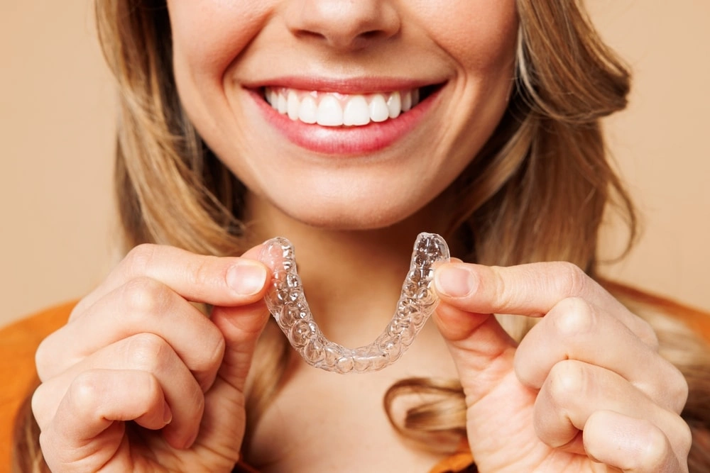 A close-up photograph showing a smiling woman with bright, aligned teeth holding a transparent orthodontic clear aligner tray directly in front of her smile. This image represents the discrete Invisalign therapies, custom clear aligner tracking systems, and comfortable aesthetic orthodontic treatments delivered at icónica dental group.