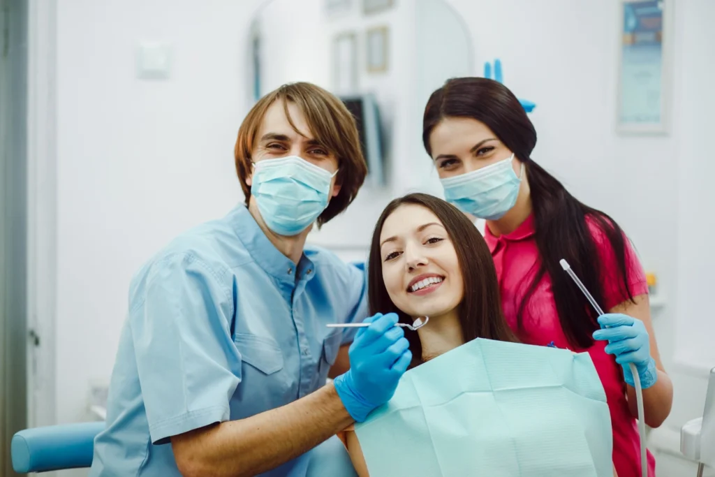 A professional clinical group photograph capturing a collaborative team care experience. A smiling female patient is seated in a treatment chair surrounded by a male clinician in blue scrubs and a female assistant in pink scrubs, both wearing surgical face masks and blue gloves while holding dental instruments. This image represents the comprehensive dental team synergy, modern clinical operatory standards, and patient-first treatment philosophy delivered at icónica dental group.