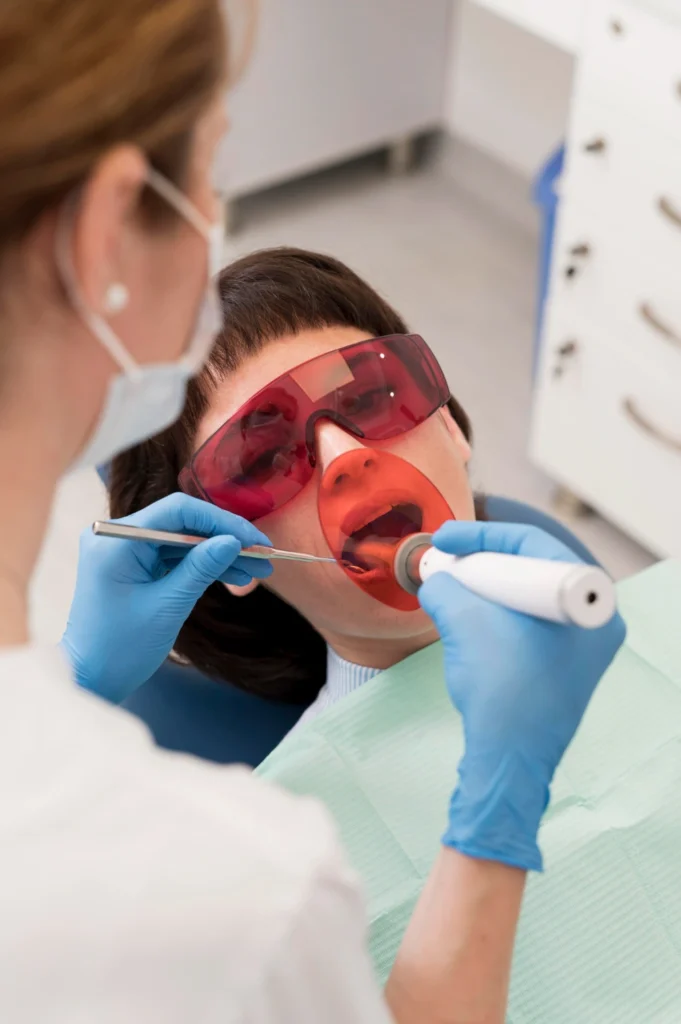 A professional photograph showing a restorative procedure in an operatory. A female patient wearing red protective eyewear and a lip retractor rests in a treatment chair while a female clinician in a white coat and blue gloves utilizes a light-curing wand to cure a resin restoration. This image represents the advanced dental bonding, high-precision composite fillings, and photo-polymerization technology delivered at icónica dental group.
