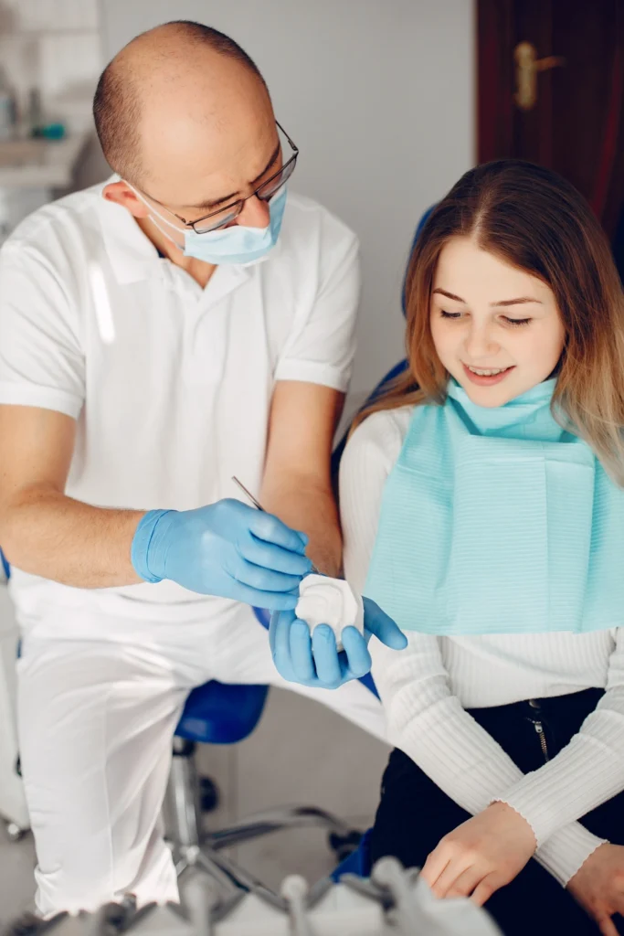 A professional photograph showing a male general dentist in glasses, a surgical mask, and blue gloves holding a dental plaster stone cast model. He is using the diagnostic model to explain a restorative or aesthetic smile alignment structure to a seated female patient wearing a teal dental bib. This image represents the detailed treatment visualization, personalized diagnostic planning, and advanced cosmetic design protocols utilized at icónica dental group.