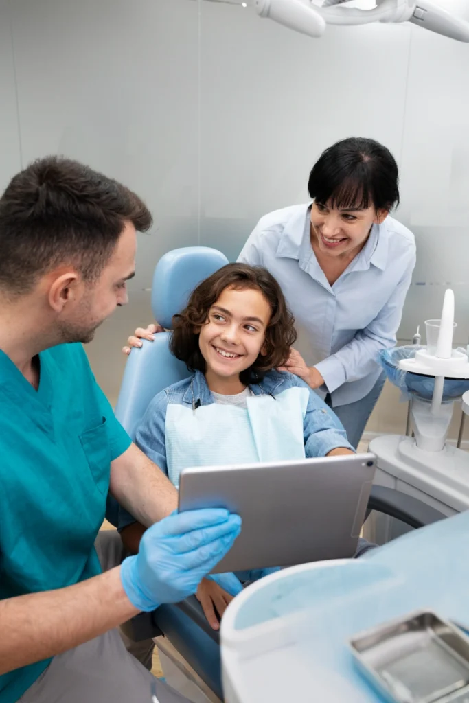 A professional photograph showing a male general dentist in teal scrubs and protective gloves holding a digital tablet while speaking with a smiling young boy in the treatment chair. A smiling woman stands supportively next to them in a bright, modern clinic. This image represents the comprehensive family dentistry, preventative care plans, and dental membership options offered at icónica dental group.