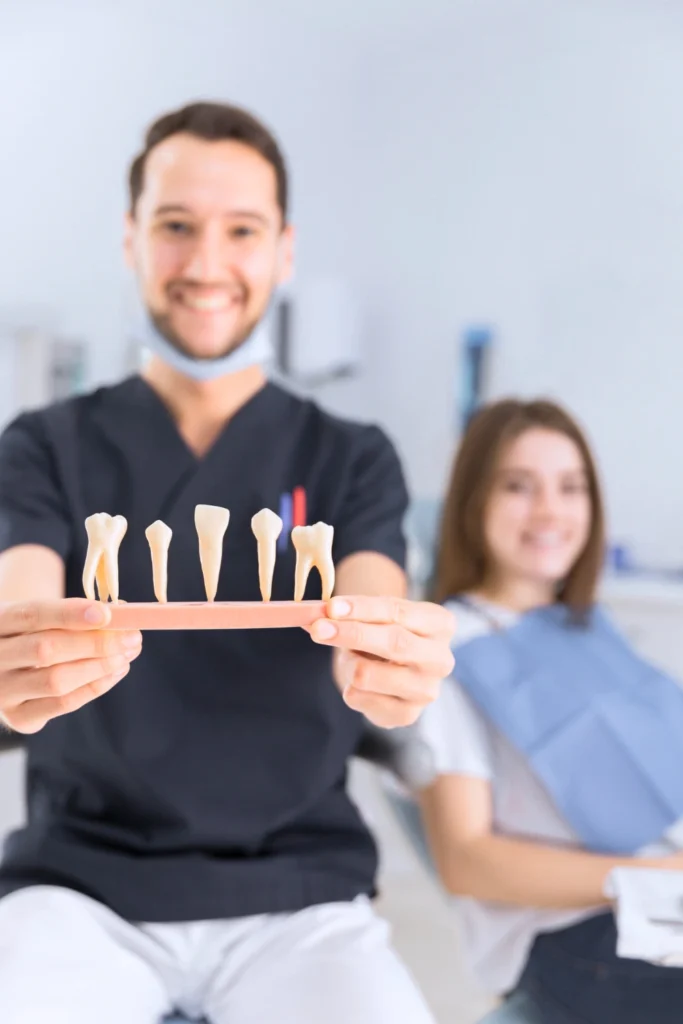 A professional photograph showing a smiling male clinician wearing dark scrubs holding a dental model block displaying individual teeth structures up toward the camera. A smiling female patient rests comfortably in the blurred background of the modern operatory chair. This image represents the advanced crown and bridge restorations, structural prosthetic mapping, and custom fixed prosthodontic options engineered at icónica dental group.