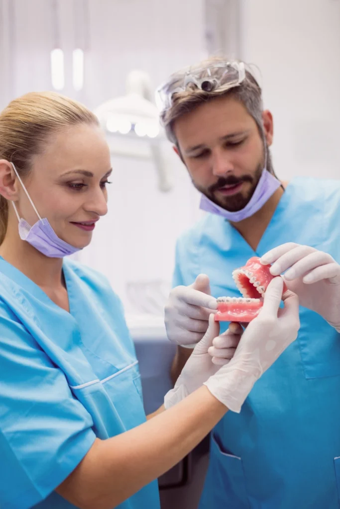 A professional photograph showing a collaborative clinical team evaluation. A male clinician wearing surgical loupes on his forehead and a female clinician, both in blue scrubs, use white protective gloves to examine an articulated complete dental model. This image represents the advanced implant-supported dentures case collaboration, detailed prosthodontic treatment mapping, and multi-disciplinary case coordination delivered at icónica dental group.