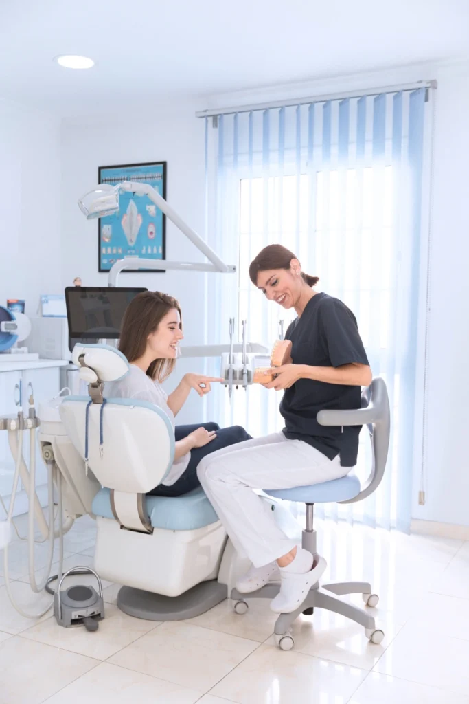A professional clinical photograph showing an interactive treatment demonstration. A smiling female clinician in dark scrubs holds an anatomical dental arch model to explain a procedure to a female patient seated in a modern dental chair. This image represents the clear patient communication, interactive case presentations, and personalized smile education delivered at icónica dental group.