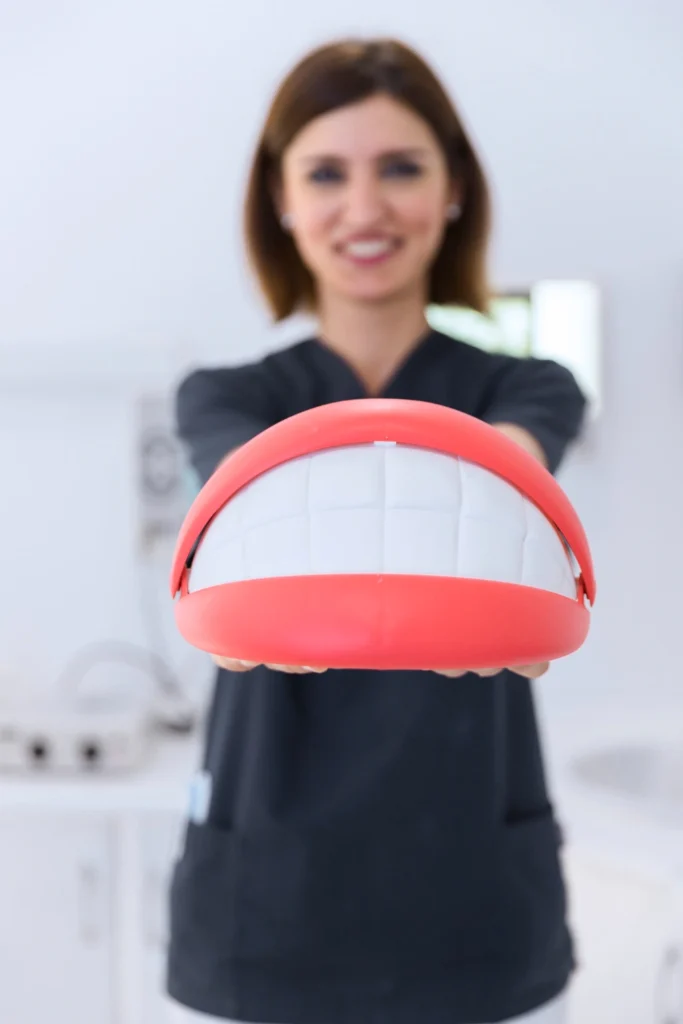 A professional photograph showing a female clinical team member in black scrubs holding a large novelty teeth and lips demonstration model out toward the camera. The clinician is smiling in a softly blurred operatory background. This image represents interactive patient education, structural smile anatomy demonstrations, and complete dental restoration guidance at icónica dental group.