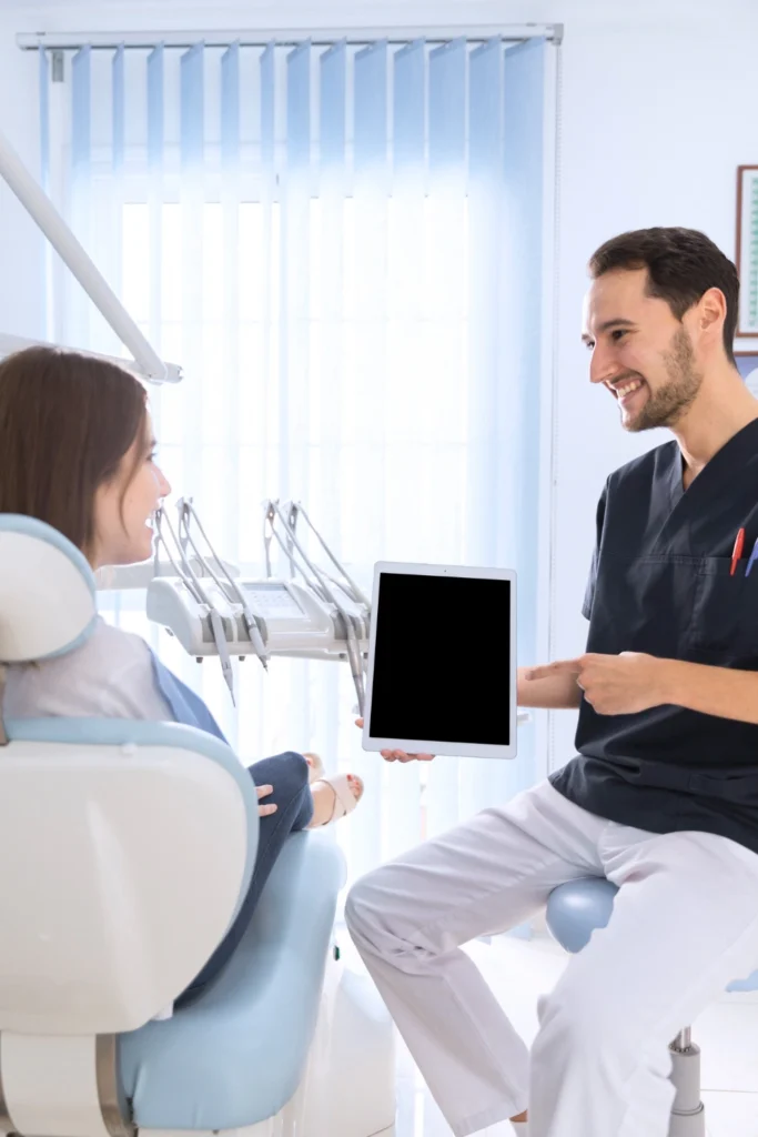 A professional clinical photograph showing a modern treatment consultation. A smiling male clinician in dark scrubs stands beside a seated female patient in a dental chair, presenting diagnostic visuals on a digital tablet computer. This image represents the transparent care consultations, interactive treatment planning, and advanced digital patient education delivered at icónica dental group.