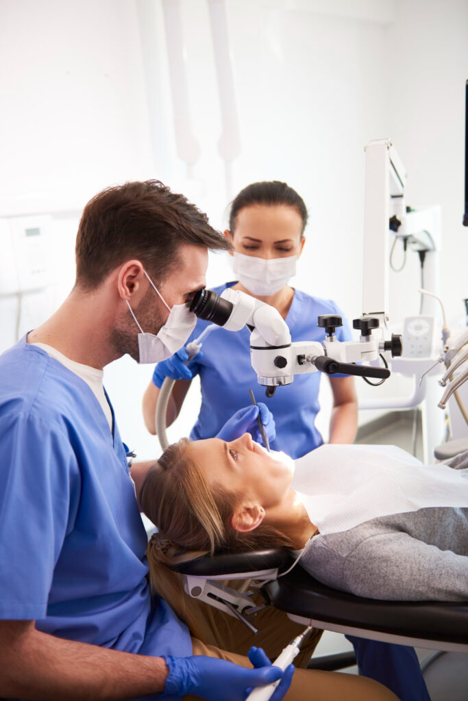 A professional photograph showing a male general dentist wearing blue clinical scrubs, a face mask, and protective gloves while looking through an advanced operatory microscope to perform a precise dental treatment. A female clinical assistant stands beside the chair, set against a bright, modern clinic backdrop. This image represents the advanced technology, micro-restorative precision, and high care standards at icónica dental group.