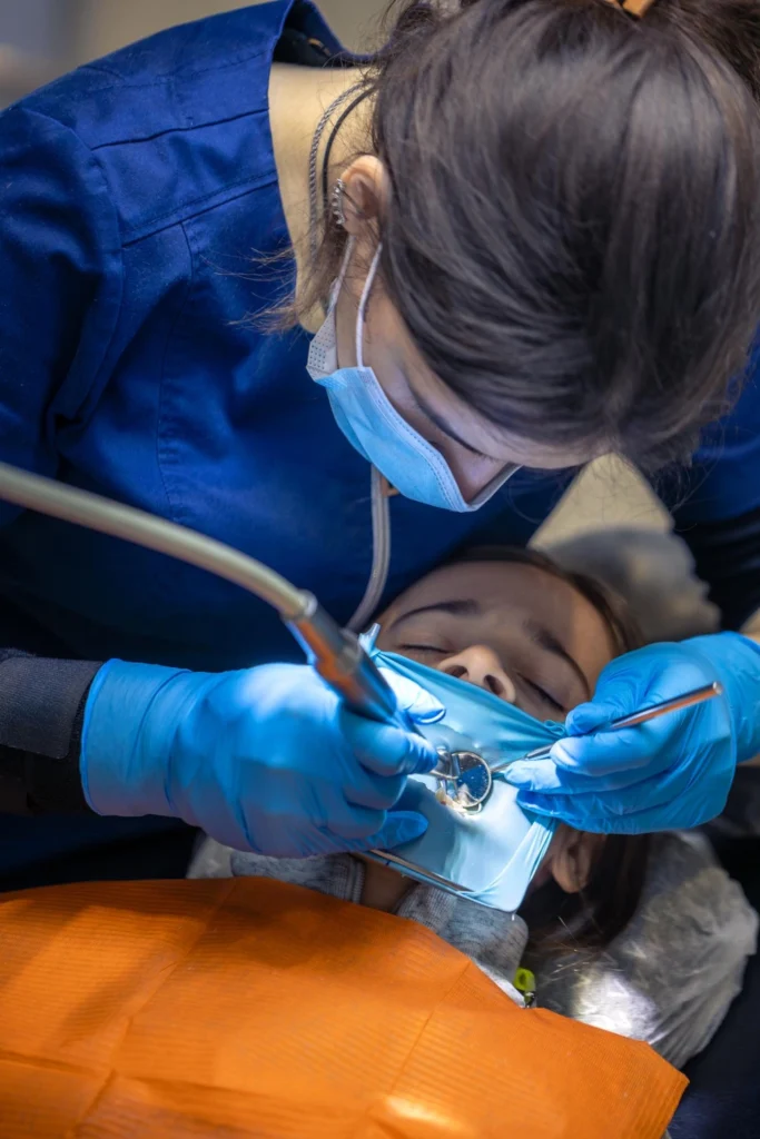 A professional close-up photograph showing an overhead view of a pediatric dental procedure. A female clinician wearing blue scrubs, a surgical mask, and blue protective gloves works with deep concentration on a young patient resting under an orange bib. The treatment site is isolated under a sterile blue rubber dam system while she utilizes a handpiece and a dental mirror tool. This image represents the precise pediatric surgical extractions, high-precision clinical isolation protocols, and specialized conservative surgery provided at icónica dental group.