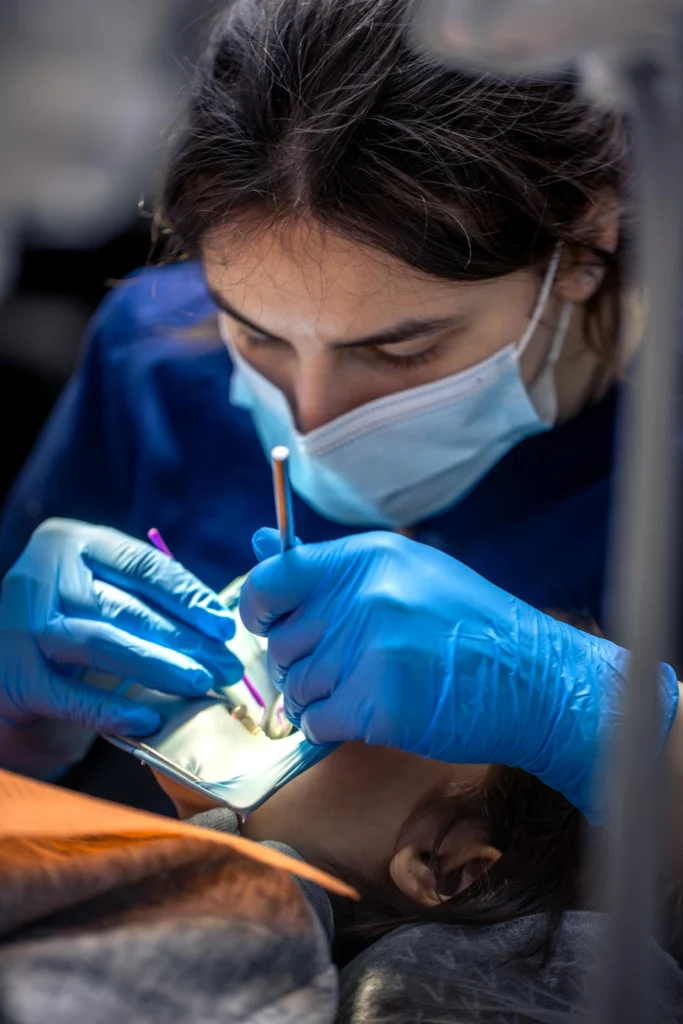 A professional close-up photograph capturing a female clinician wearing a surgical face mask and blue protective gloves as she performs a precise pre-prosthetic surgical procedure. The treatment site is isolated under a clinical teal rubber dam system to ensure optimal sterility and view. This image represents the advanced clinical isolation protocols, meticulous surgical execution, and precise pre-prosthetic care delivered at icónica dental group.