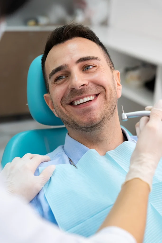 A professional photograph showing a relaxed, smiling male patient resting comfortably in a teal dental chair with a light blue bib. A clinician wearing white protective gloves holds a precise clinical handpiece instrument nearby. This image represents the high-end patient comfort standards, elite preventative dental care, and reassuring clinical experiences delivered at icónica dental group.