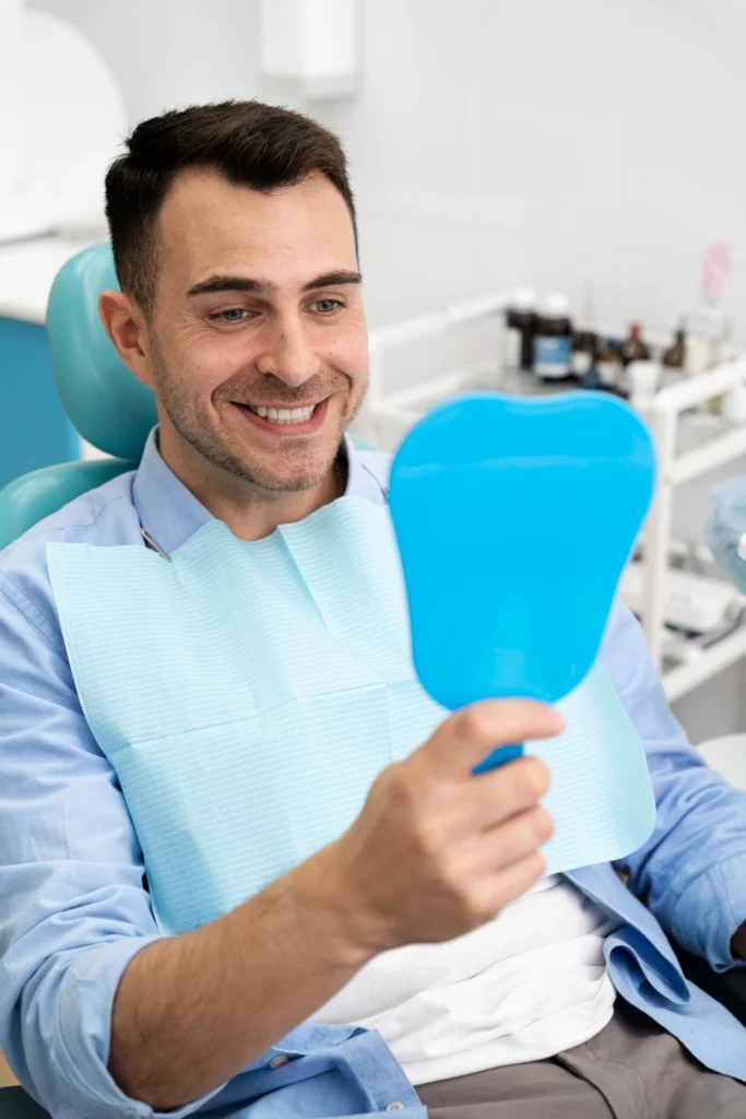 A professional photograph showing a happy, smiling male patient resting in a dental chair with a blue bib. He holds up a tooth-shaped hand mirror to happily admire his restored smile. This image represents the single tooth implant restorations, seamless final crown placements, and life-changing aesthetic transformations delivered at icónica dental group.