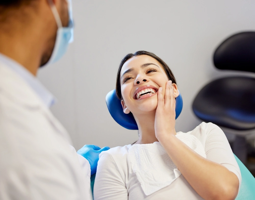 A professional photograph showing a happy, smiling female patient resting in a dental chair during a post-operative evaluation. She gently touches her cheek while consulting with a male clinician wearing a white lab coat and protective face mask. This image represents the successful post-surgical recoveries, compassionate clinical support, and personalized bone grafting follow-up care provided at icónica dental group.