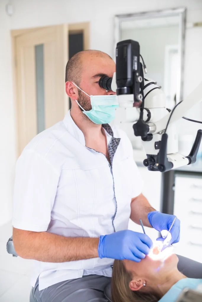 A professional photograph showing a male general dentist in a white clinic jacket, a surgical mask, and blue gloves looking through an advanced clinical operatory microscope. He is executing a highly focused restorative procedure on a seated patient. This image represents the advanced surgical microscopy, micro-endodontic technology, and conservative tooth-saving standards implemented at icónica dental group.