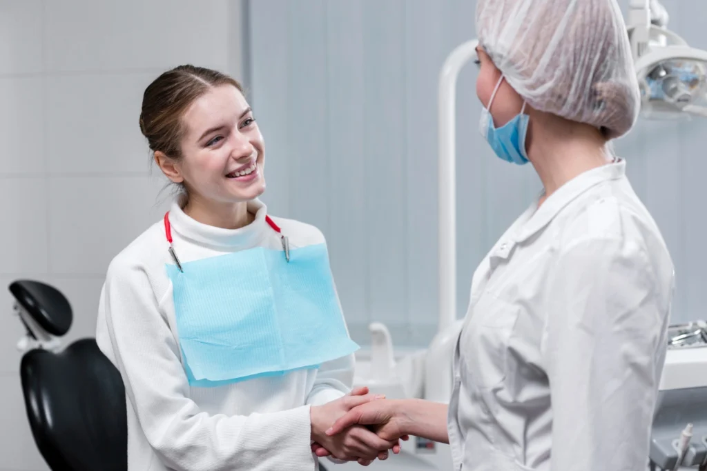 A professional clinical photograph showing a warm patient greeting. A smiling female patient wearing a blue dental bib stands and shakes hands with a female clinician wearing a white lab coat, protective hairnet, and mask inside a modern treatment operatory. This image represents the welcoming patient onboarding experiences, professional relationship building, and foundational trust delivered at icónica dental group.