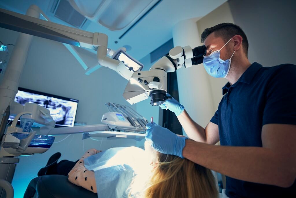 A professional photograph of a male general dentist wearing a face mask, protective gloves, and clinical scrubs, operating an advanced dental microscope under cool operatory lighting. He is performing a highly precise root canal therapy or micro-restorative procedure on a patient in the chair. This image represents the advanced technology and diagnostic precision utilized to provide comfortable endodontic treatment at icónica dental group.