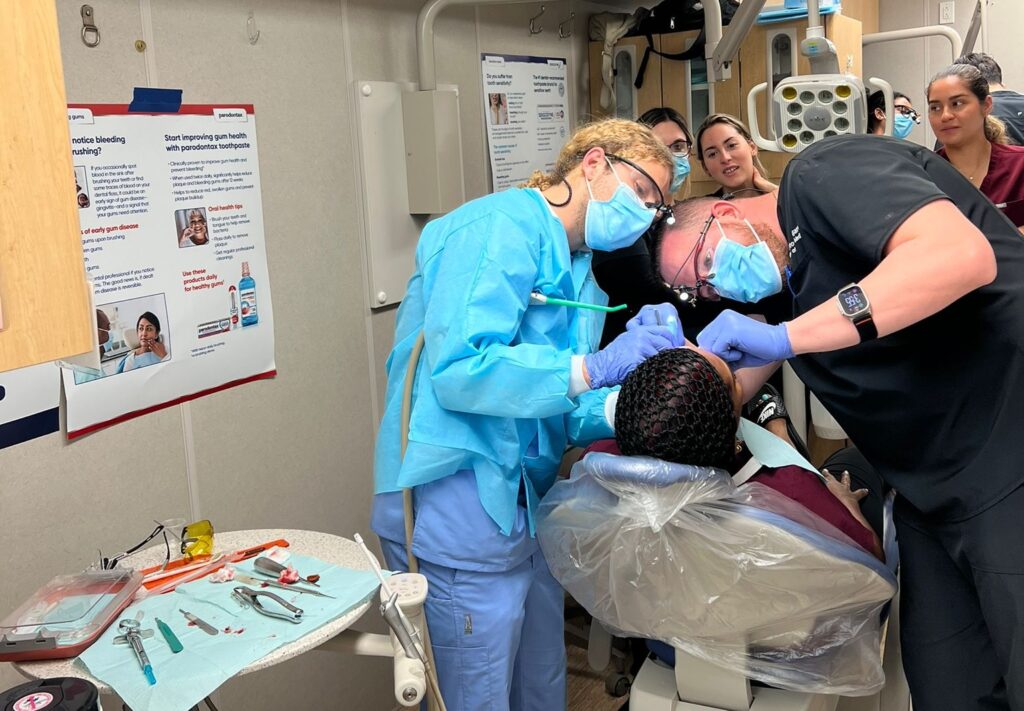 An authentic clinical photograph showing Dr. Christopher Garcia and his surgical outreach team wearing blue protective gowns, surgical face masks, and loupes while performing a dental procedure on a patient in a clinic chair. Other international clinical students observe in the background. This image highlights the international dental training, humanitarian mission experience, and global outreach behind the patient care standards at icónica dental group.