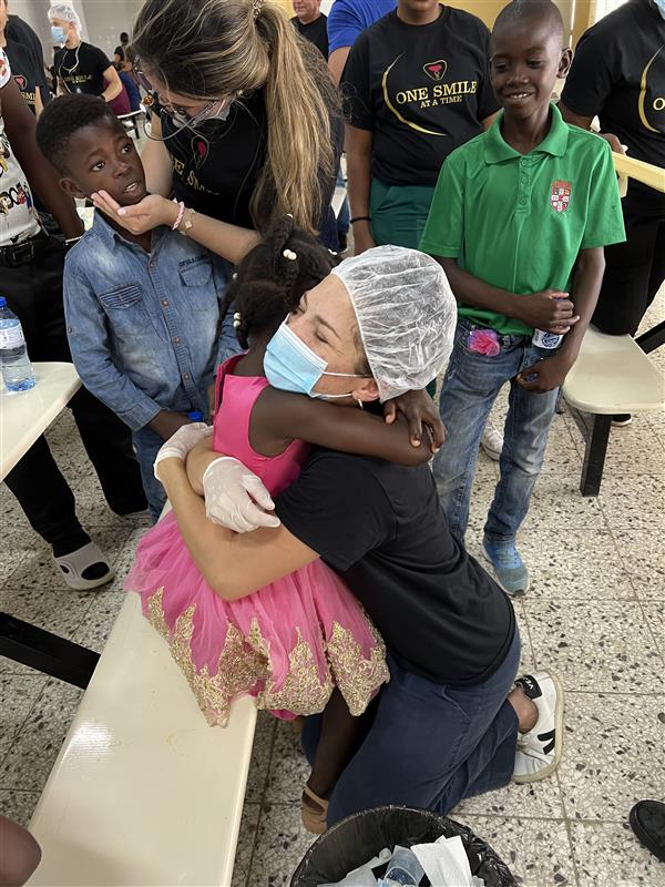 An authentic outreach photograph from a humanitarian dental mission. A female team member wearing a clinical hair net, face mask, and protective gloves kneels down to embrace a young girl in a pink dress, while local children and volunteers in 'One Smile at a Time' t-shirts smile in the background. This image highlights the international dental outreach, compassionate pediatric care, and global philanthropy foundational to the mission of icónica dental group.