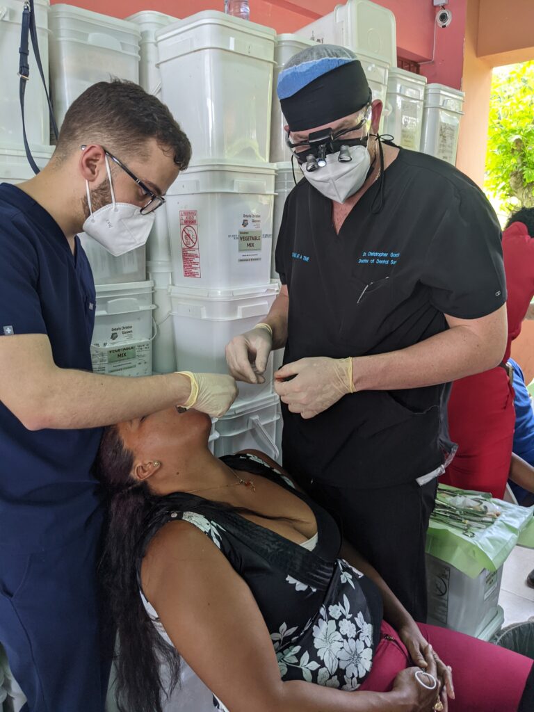 An authentic clinical photograph showing Dr. Christopher Garcia in black scrubs and surgical loupes working with a clinical assistant in blue scrubs and an N95 mask. They are performing a pro bono dental surgery for a patient in a chair during a One Smile at a Time non-profit humanitarian mission. This image represents the advanced oral surgery skills, clinical field training, and community philanthropy behind the care at icónica dental group.