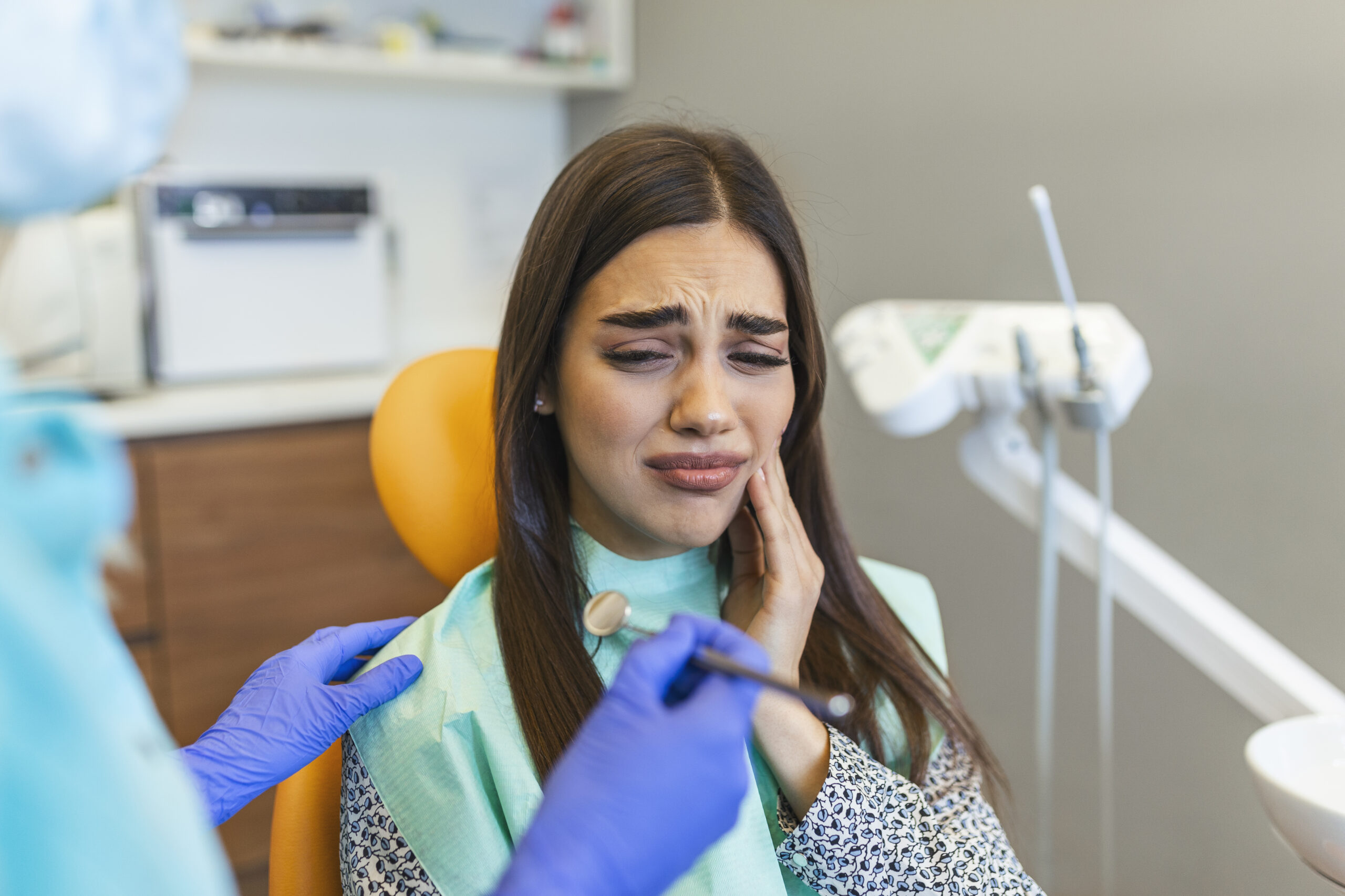 A photograph showing a young woman sitting in a dental treatment chair, holding her hand to her cheek with an expression of severe pain from a toothache. A clinician wearing blue protective gloves gently comforts her during an urgent diagnostic evaluation. This image represents the compassionate emergency dental care and immediate pain relief protocols provided at icónica dental group.