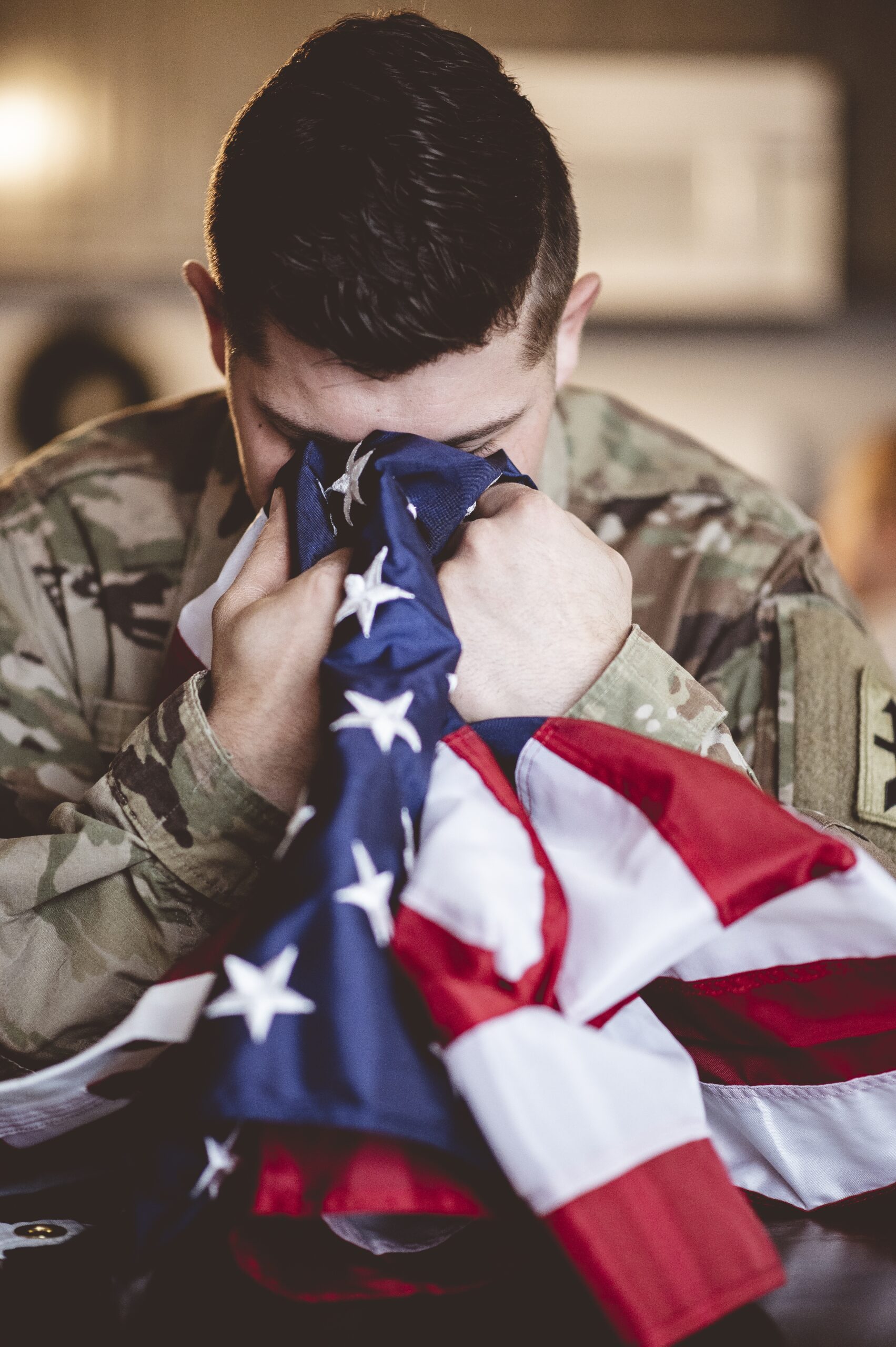 A poignant vertical photograph of a military service member in camouflage uniform, holding a folded American flag close to his face in a moment of deep reflection or prayer. This image represents the profound sacrifice of our military service members and the deep commitment of the 'One Smile at a Time' program to provide pro bono dental care to veterans at icónica dental group.