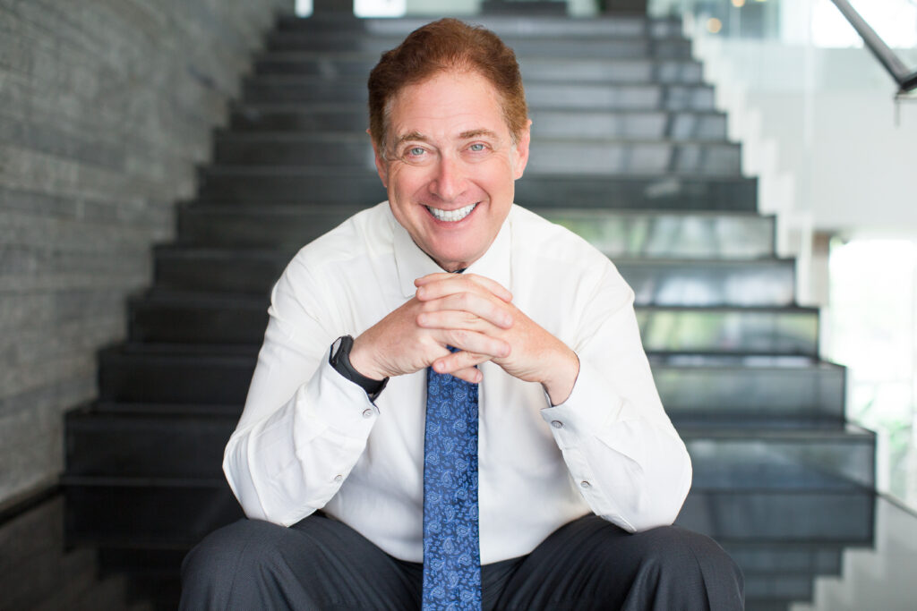 A professional portrait focusing on a smiling executive sitting forward with hands clasped, wearing a white dress shirt and a blue patterned tie against a modern blurred stone staircase backdrop. This image represents the comprehensive oral health optimization and custom smile design treatments tailored for corporate professionals at icónica dental group.
