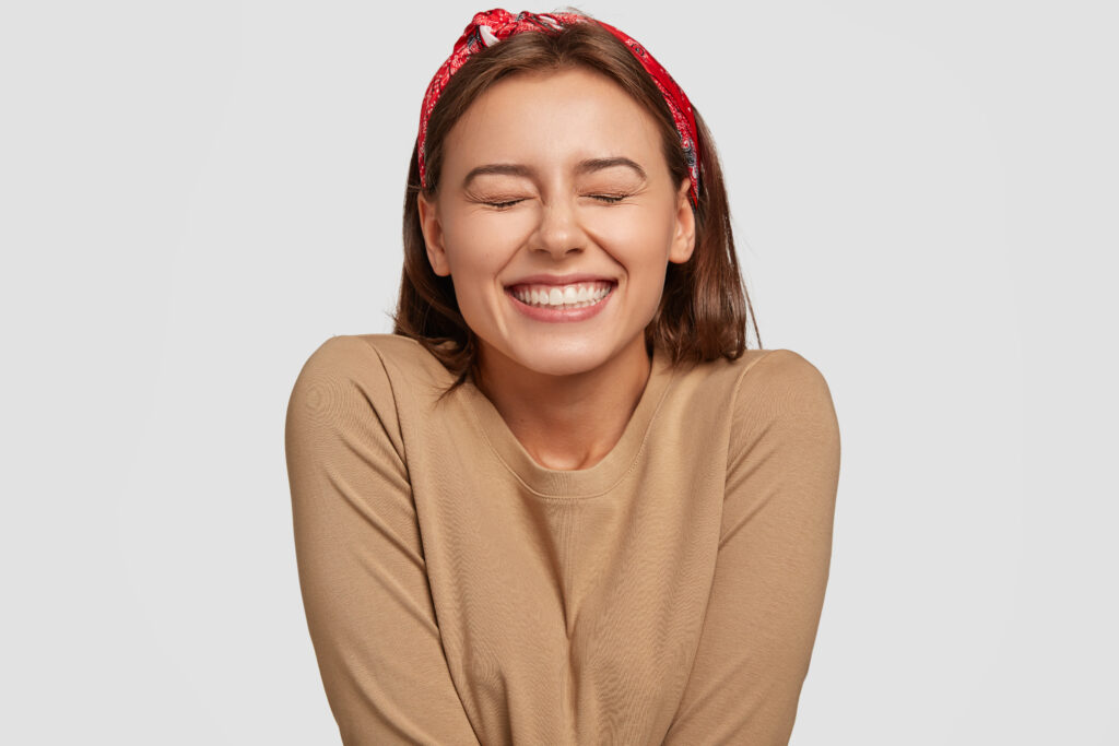 A studio portrait focusing on a woman with a vibrant, beautifully aligned, and radiant broad smile. She has her eyes closed in joy and is wearing a red patterned headband with a tan sweater against a neutral grey background. This image represents the life-changing aesthetic transformations and premium porcelain veneer treatments designed to deliver absolute confidence at icónica dental group.